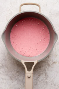 A beige saucepan filled with a pink liquid mixture sits on a light textured surface, viewed from above.