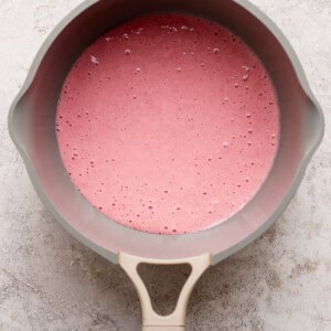 A beige saucepan filled with a pink liquid mixture sits on a light textured surface, viewed from above.