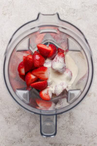 Overhead view of a blender containing halved strawberries and a scoop of white powder on a light textured surface.