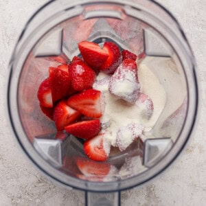 Overhead view of a blender containing halved strawberries and a scoop of white powder on a light textured surface.