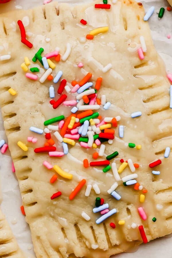 A rectangular pastry topped with icing and colorful sprinkles sits on parchment paper next to a fresh strawberry.