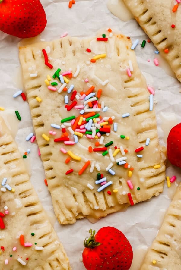 Rectangular pastry with icing and colorful sprinkles, surrounded by fresh strawberries on a sheet of parchment paper.