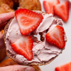 A hand holds a bagel half topped with pink cream cheese and sliced strawberries, with more strawberries and bagels on a plate in the background.