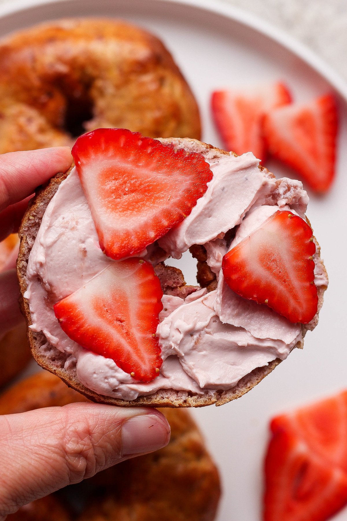 A hand holds a bagel half topped with pink cream cheese and sliced strawberries, with more strawberries and bagels on a plate in the background.