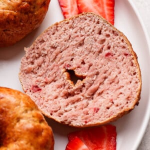 A sliced strawberry bagel is shown on a white plate with two fresh strawberry slices underneath.