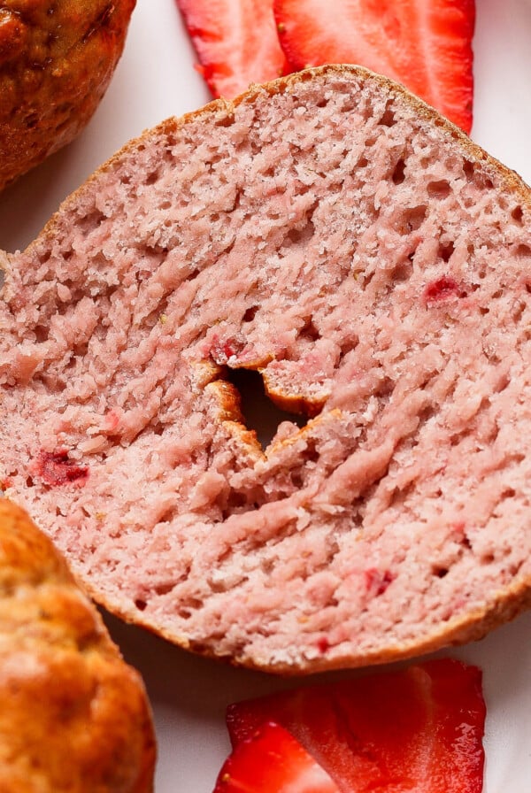 A sliced strawberry bagel is shown on a white plate with two fresh strawberry slices underneath.