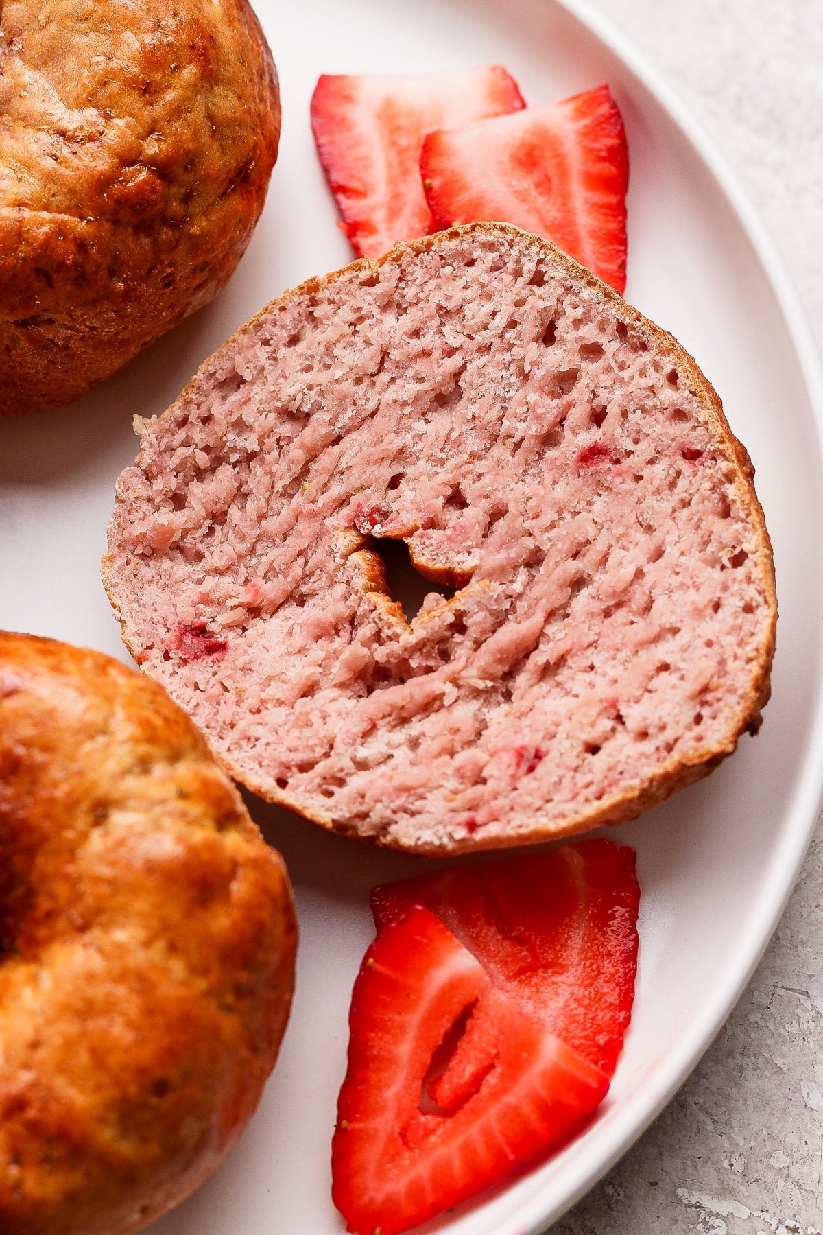 A sliced strawberry bagel on a plate with three fresh strawberry slices, next to a whole bagel.