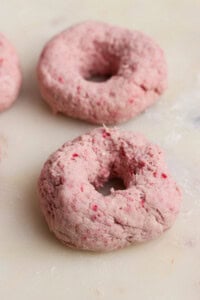 Two unbaked, pinkish doughnuts made with a textured dough rest on a light-colored surface.