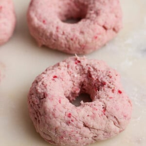 Two unbaked, pinkish doughnuts made with a textured dough rest on a light-colored surface.