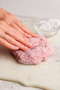 A person kneads pink dough on a floured surface with a bowl of flour in the background.