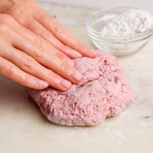 A person kneads pink dough on a floured surface with a bowl of flour in the background.
