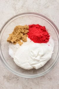 A glass bowl containing flour, brown sugar, white sugar, red powder, and a dollop of yogurt on a light surface.