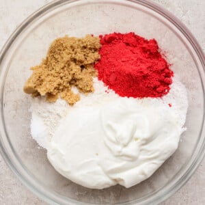 A glass bowl containing flour, brown sugar, white sugar, red powder, and a dollop of yogurt on a light surface.