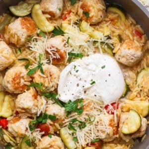 A skillet filled with orzo pasta, zucchini, meatballs, diced tomatoes, shredded cheese, a dollop of sour cream, and chopped parsley.