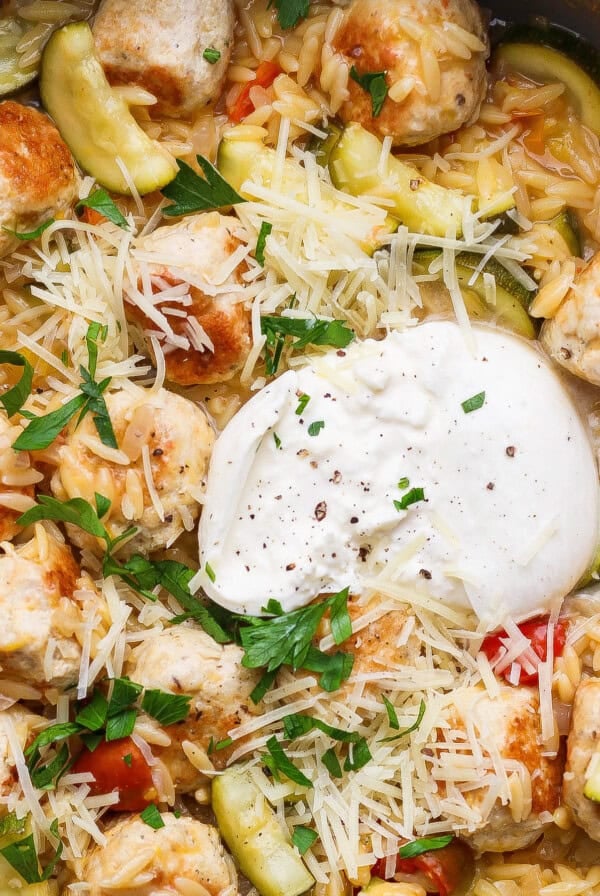 A skillet filled with orzo pasta, zucchini, meatballs, diced tomatoes, shredded cheese, a dollop of sour cream, and chopped parsley.