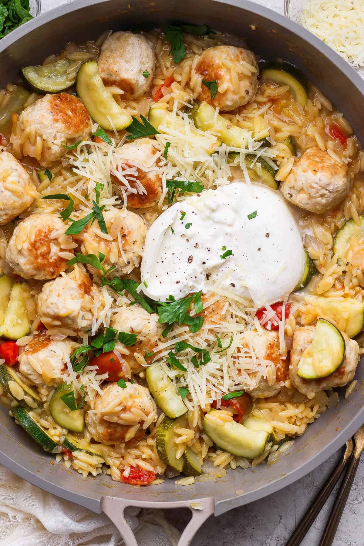 A skillet filled with orzo pasta, chicken meatballs, sliced zucchini, red peppers, grated cheese, fresh parsley, and a dollop of burrata cheese in the center.