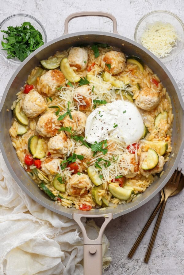 A skillet with chicken meatballs, orzo pasta, zucchini, cherry tomatoes, shredded cheese, and a dollop of burrata, garnished with herbs. Two small bowls with cheese and herbs are nearby.