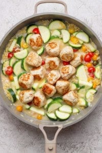 A skillet with browned meatballs, sliced zucchini, yellow squash, cherry tomatoes, and orzo in a light sauce on a gray surface.