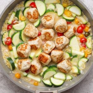 A skillet with browned meatballs, sliced zucchini, yellow squash, cherry tomatoes, and orzo in a light sauce on a gray surface.