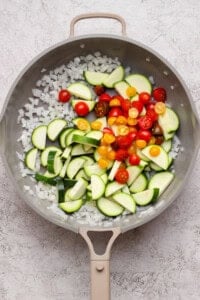 A frying pan contains chopped onions, sliced zucchini, and halved cherry tomatoes on a light textured surface.