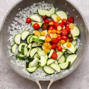 A frying pan contains chopped onions, sliced zucchini, and halved cherry tomatoes on a light textured surface.