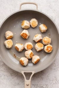 A beige skillet contains partially cooked, lightly browned meatballs spread out on its surface.