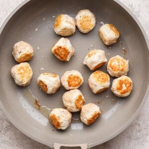 A beige skillet contains partially cooked, lightly browned meatballs spread out on its surface.