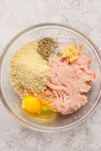 A glass bowl containing ground chicken, a cracked egg, breadcrumbs, minced garlic, salt, and dried herbs on a light textured surface.