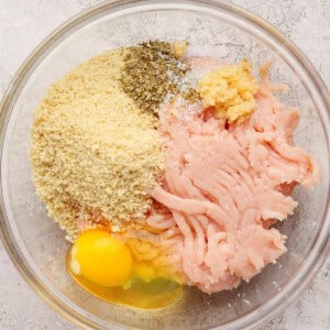 A glass bowl containing ground chicken, a cracked egg, breadcrumbs, minced garlic, salt, and dried herbs on a light textured surface.