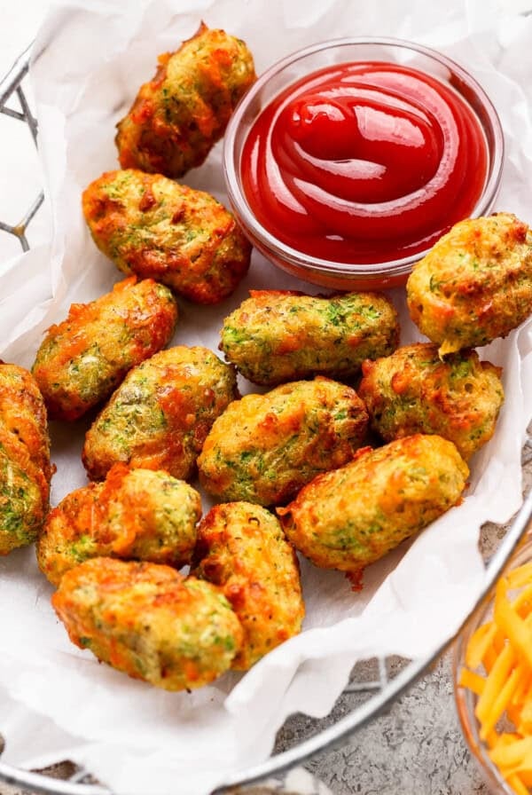 A basket of golden-brown vegetable tots on parchment paper, served with a small glass bowl of ketchup for dipping.