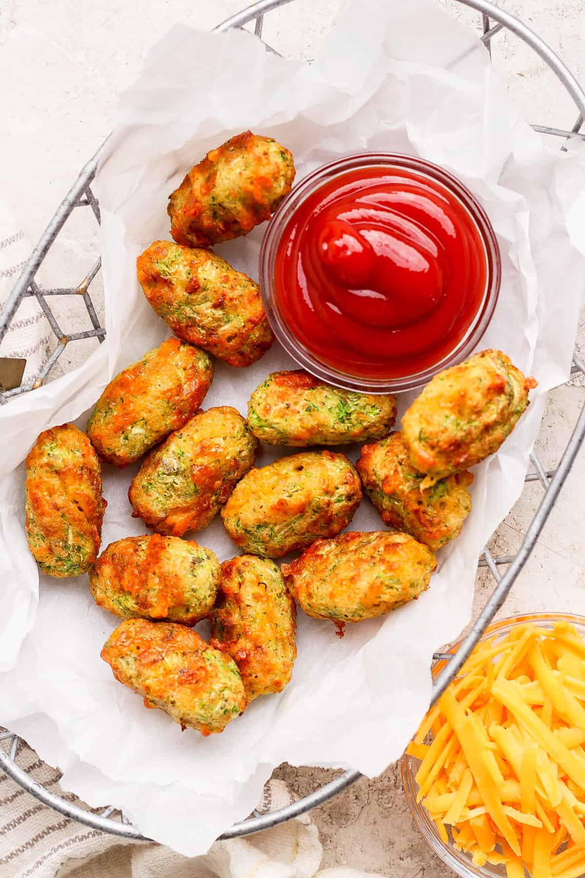 A basket of baked vegetable tots on parchment paper served with a bowl of ketchup; shredded cheese is visible beside the basket.