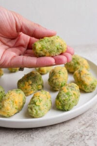 A hand holds a homemade broccoli and cheese tot over a white plate with more tots arranged on it.