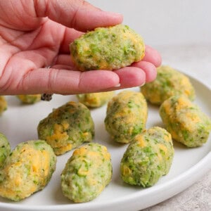 A hand holds a homemade broccoli and cheese tot over a white plate with more tots arranged on it.