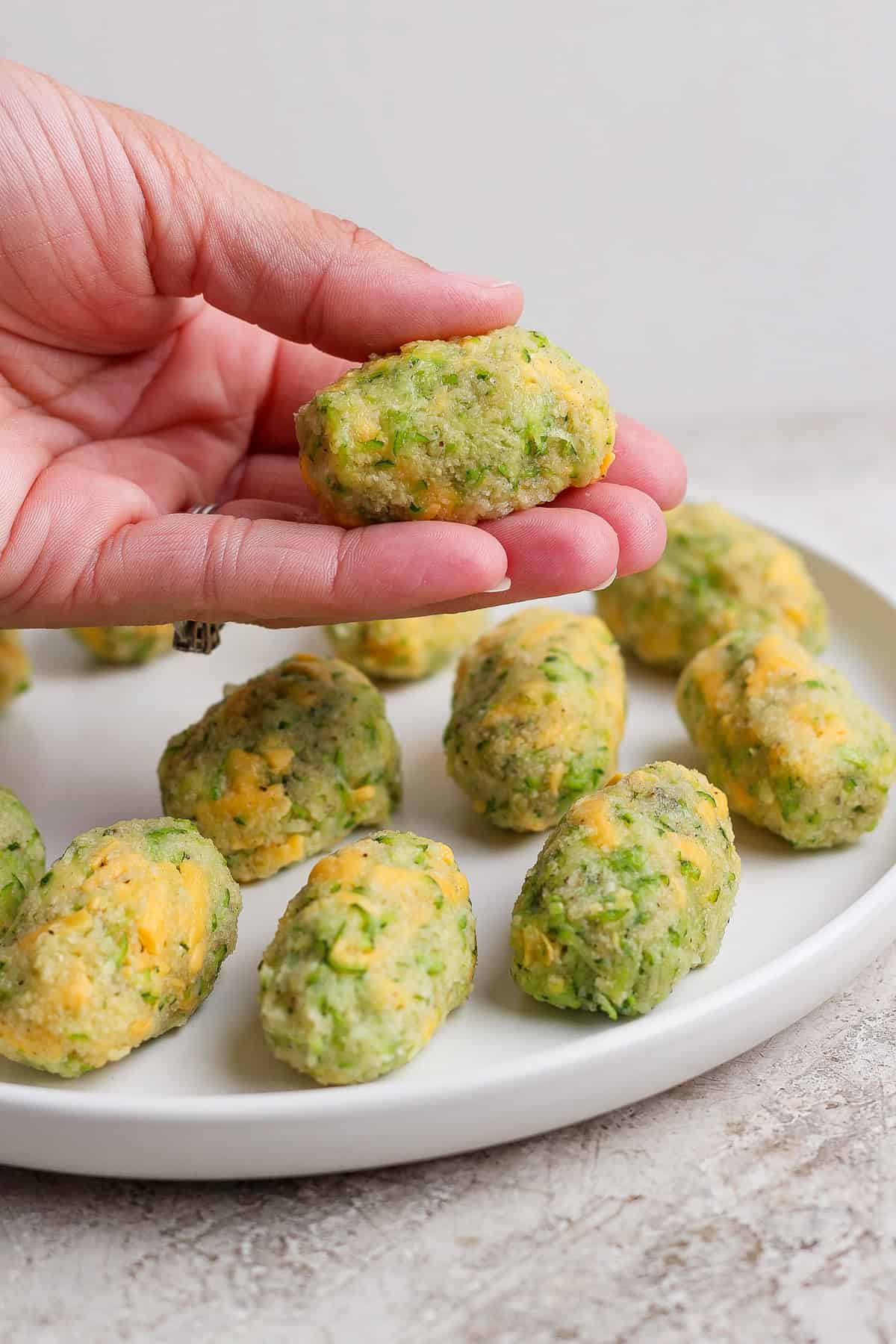 A hand holds a homemade broccoli and cheese tot over a white plate with more tots arranged on it.