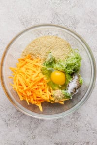 A glass bowl containing shredded cheddar cheese, grated zucchini, an egg, breadcrumbs, and seasonings on a light textured surface.