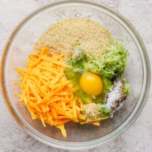 A glass bowl containing shredded cheddar cheese, grated zucchini, an egg, breadcrumbs, and seasonings on a light textured surface.