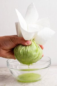 A hand squeezes shredded green zucchini wrapped in paper towels over a glass bowl, extracting liquid.