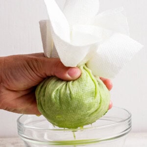 A hand squeezes shredded green zucchini wrapped in paper towels over a glass bowl, extracting liquid.