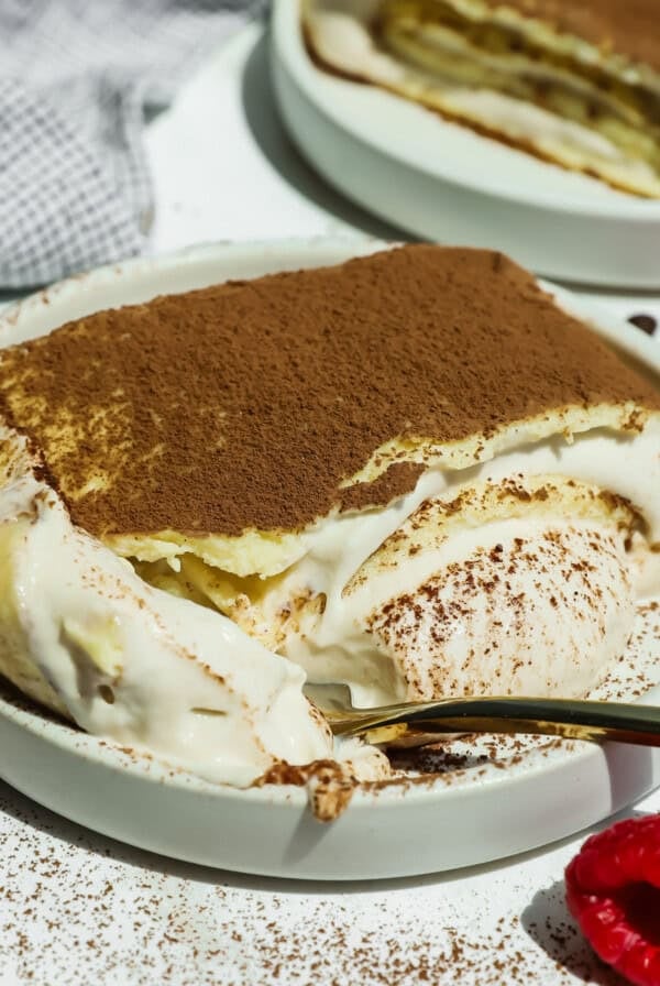 A plate of cottage cheese tiramisu crepe roll with a spoonful taken out, topped with cocoa powder, sits next to fresh raspberries on a white surface.
