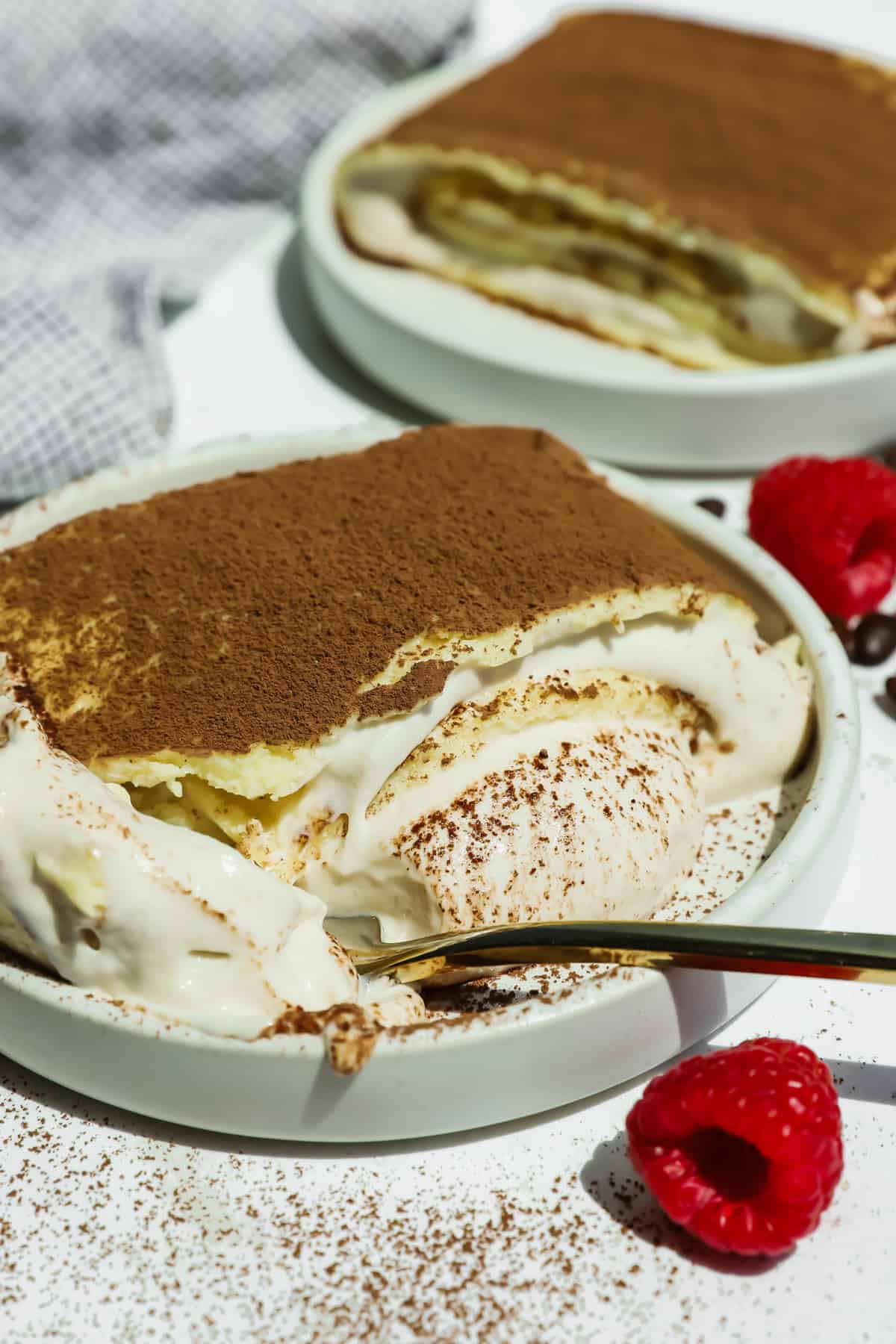 A plate of cottage cheese tiramisu crepe roll with a portion scooped out reveals creamy layers, dusted with cocoa powder. Fresh raspberries and a gold spoon are nearby.