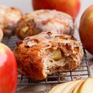 Close-up of a glazed apple pastry, reminiscent of air fryer apple fritters, with a bite taken out, resting on a cooling rack and surrounded by fresh apples and apple slices.
