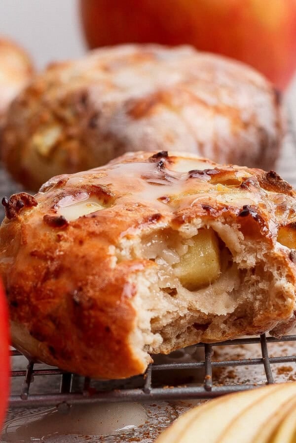 Close-up of a glazed apple pastry, reminiscent of air fryer apple fritters, with a bite taken out, resting on a cooling rack and surrounded by fresh apples and apple slices.