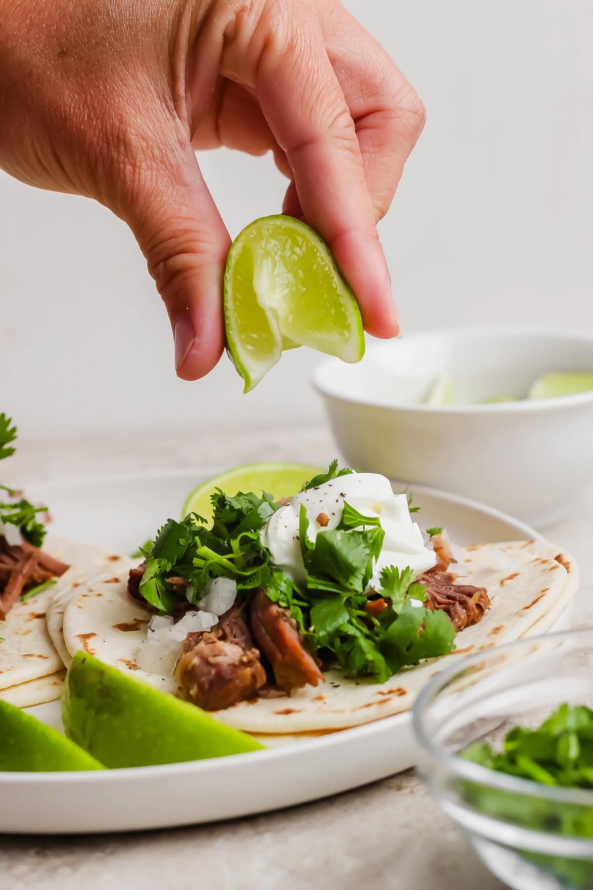 A hand squeezes a lime wedge over a taco filled with meat, cilantro, and sour cream on a white plate. Other lime wedges and a bowl are in the background.