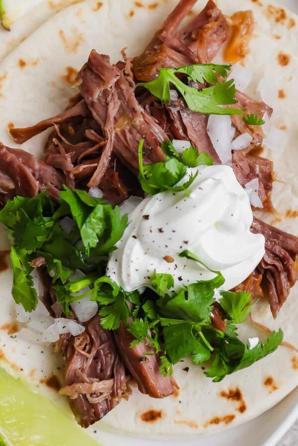 A flour tortilla topped with shredded beef, chopped onions, fresh cilantro, and a dollop of sour cream, served with a lime wedge.