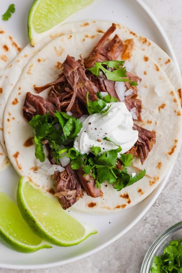 A soft flour tortilla topped with shredded meat, chopped onions, cilantro, and a dollop of sour cream, served with lime wedges on a white plate.