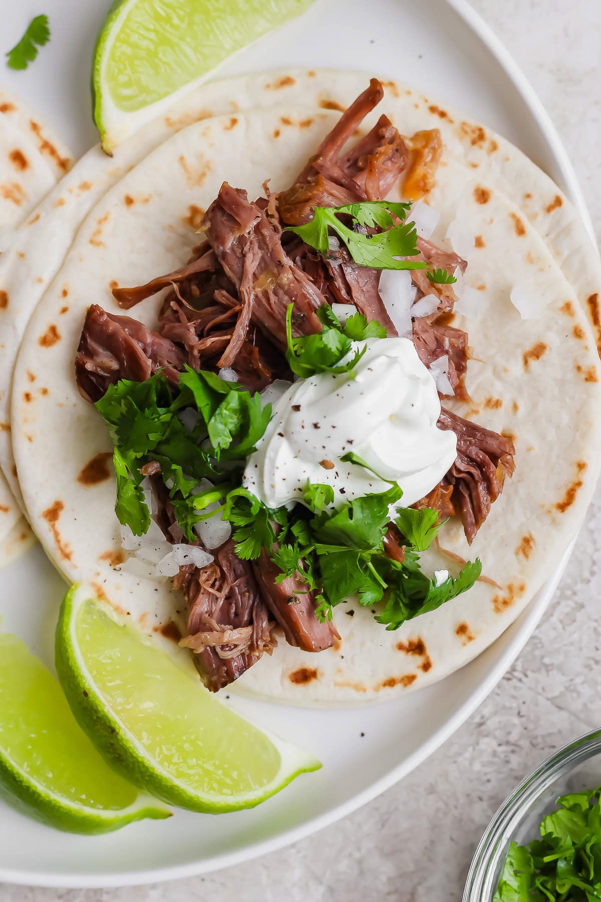 A soft flour tortilla topped with shredded meat, chopped onions, cilantro, and a dollop of sour cream, served with lime wedges on a white plate.