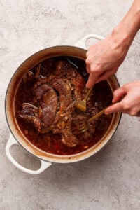 A person uses two forks to shred cooked beef in a round, white Dutch oven filled with a rich, brown sauce.