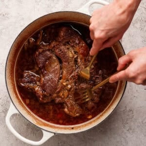 A person uses two forks to shred cooked beef in a round, white Dutch oven filled with a rich, brown sauce.