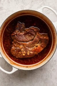 A braised beef roast sits in a white Dutch oven with a rich, red-brown broth and diced onions on a light textured surface.