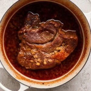 A braised beef roast sits in a white Dutch oven with a rich, red-brown broth and diced onions on a light textured surface.
